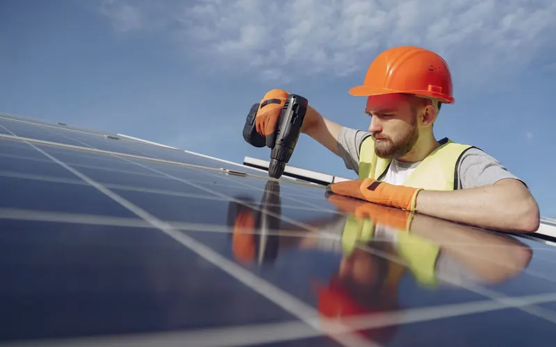 Electrician in a hard hat fixing a solar panel to its mounting rail with a drill